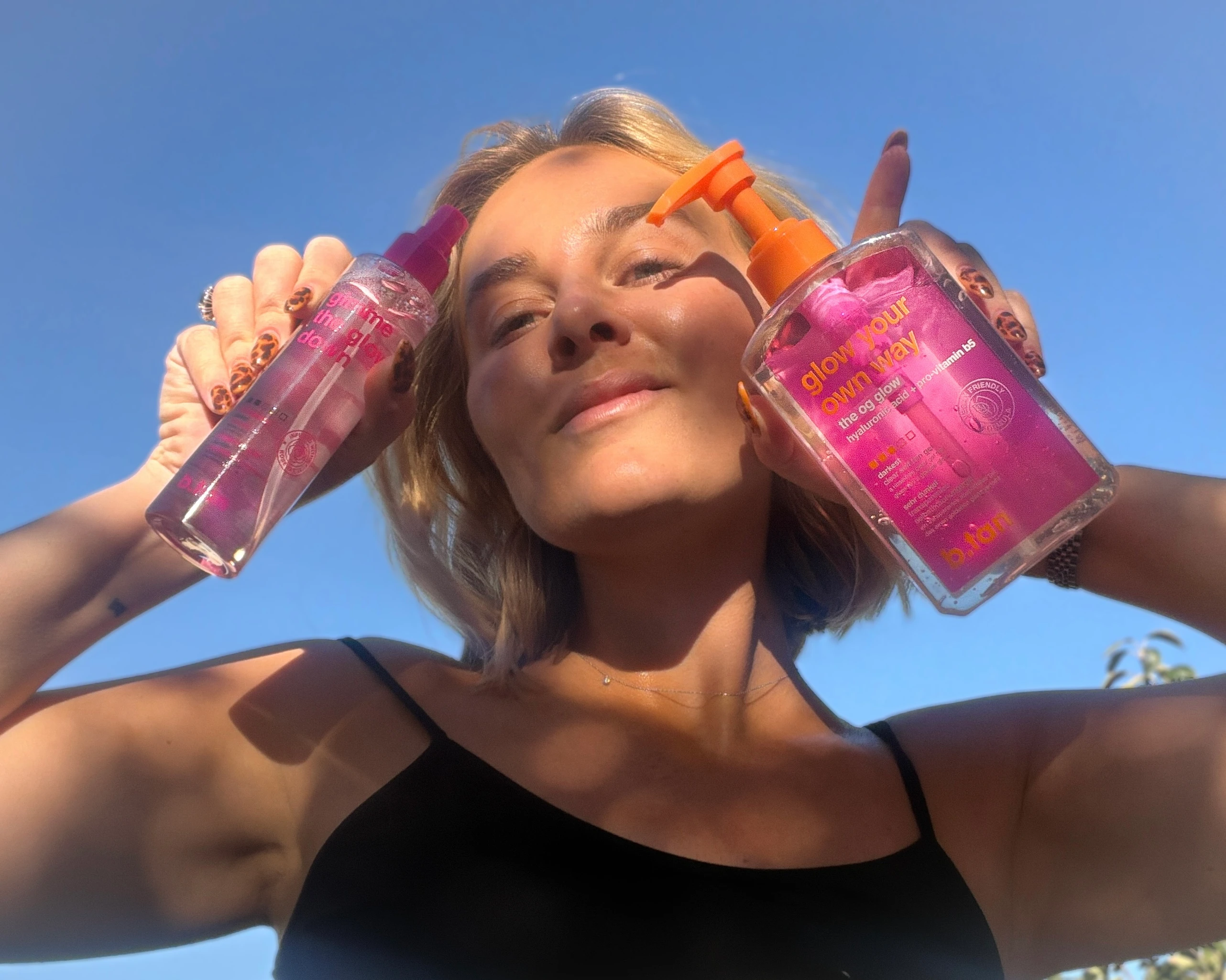 En person holder to pink b.tan 'Glow Your Own Way' og 'Glow me the glow down' hudplejeprodukter op mod en blå himmel.
