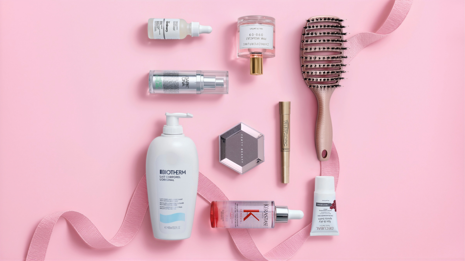 Forskellige skønhedsprodukter: hudpleje, makeup, parfume, hårbørste og lotion, arrangeret på lyserød baggrund med bånd.