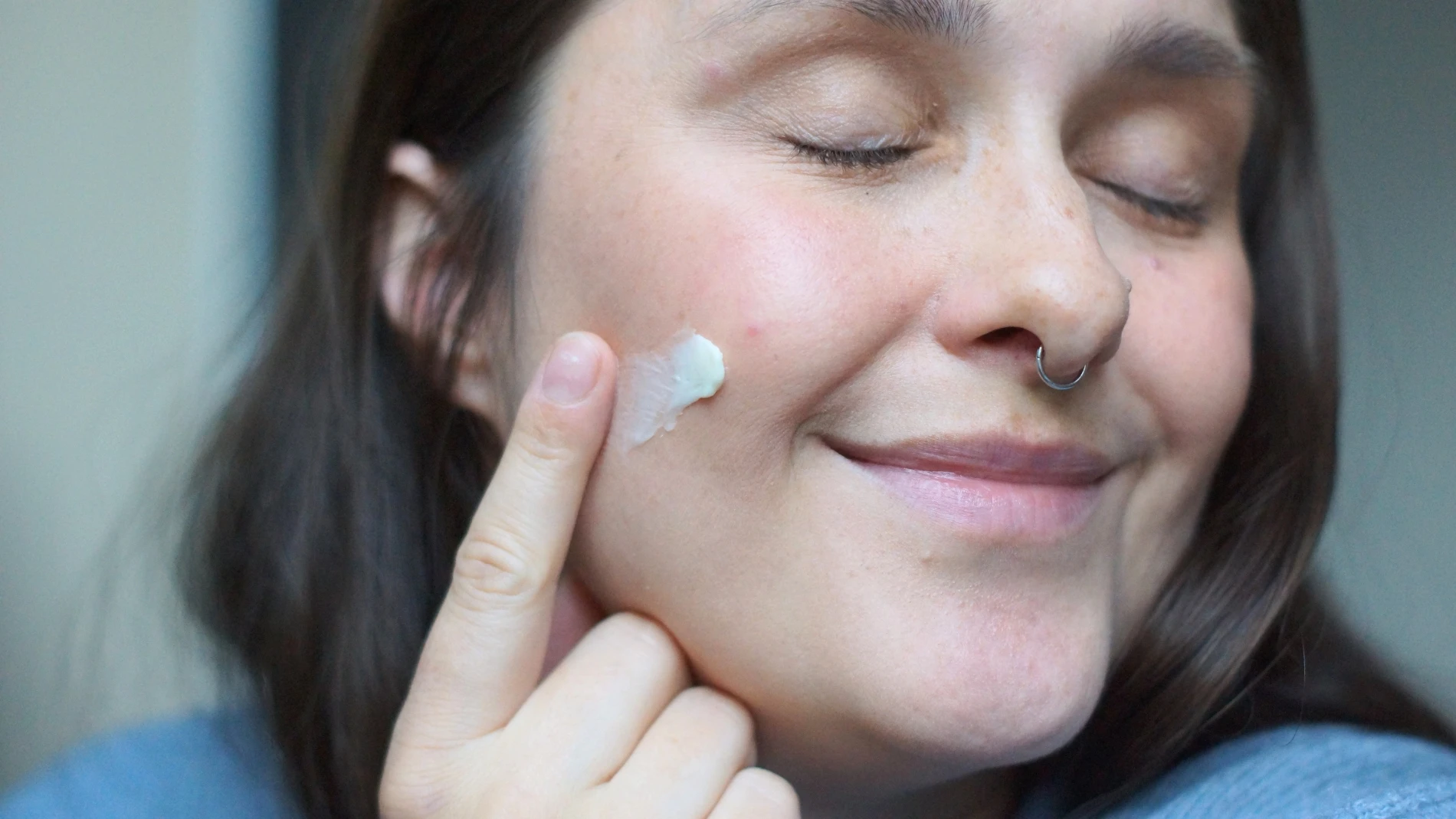 En person med lukkede øjne og et let smil påfører en grønlig creme på sin kind med en finger.