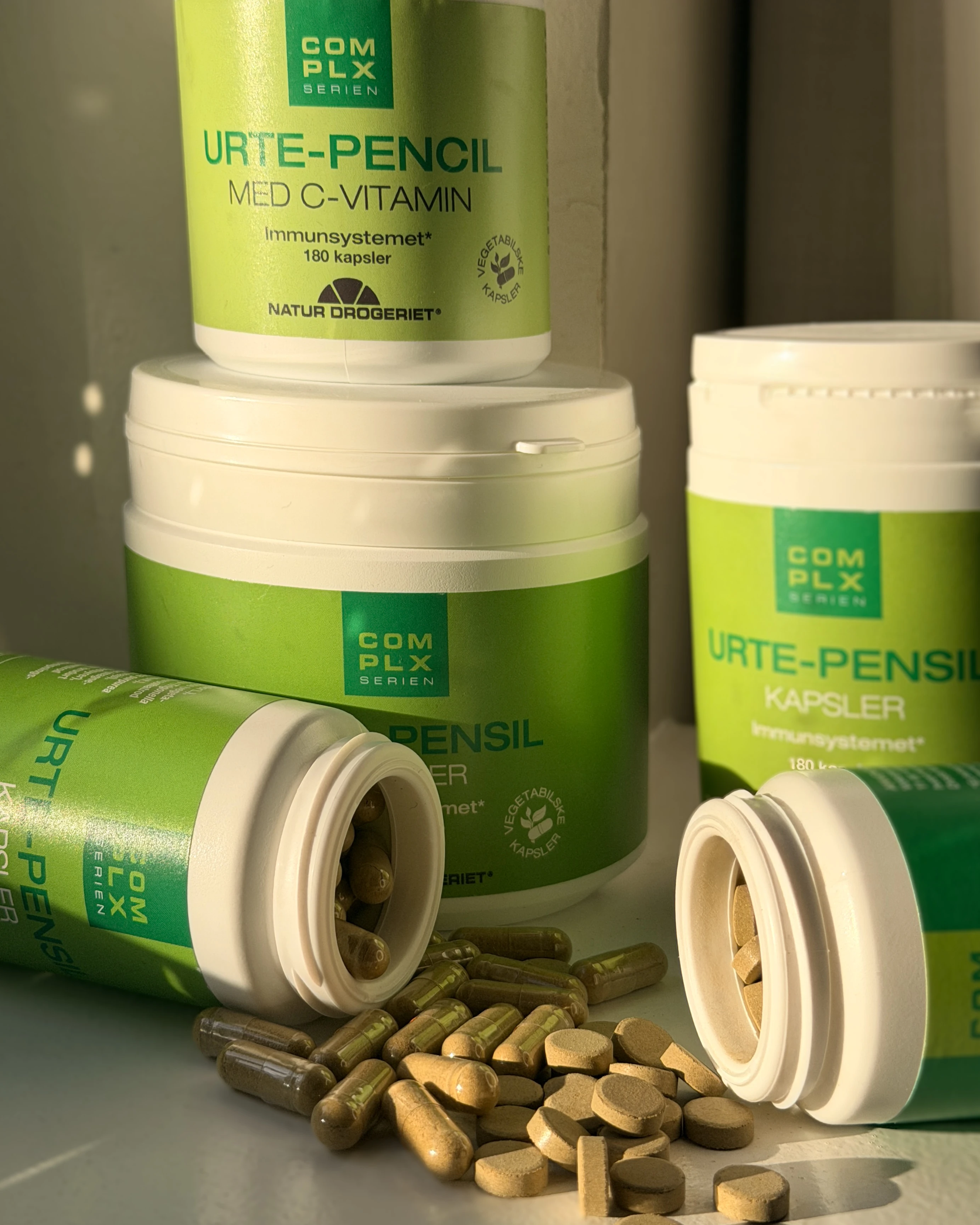 Grønne beholdere af Natur Drogeriet Urte-Pensil med C-vitamin. En er væltet, med udspredte kapsler og tabletter.