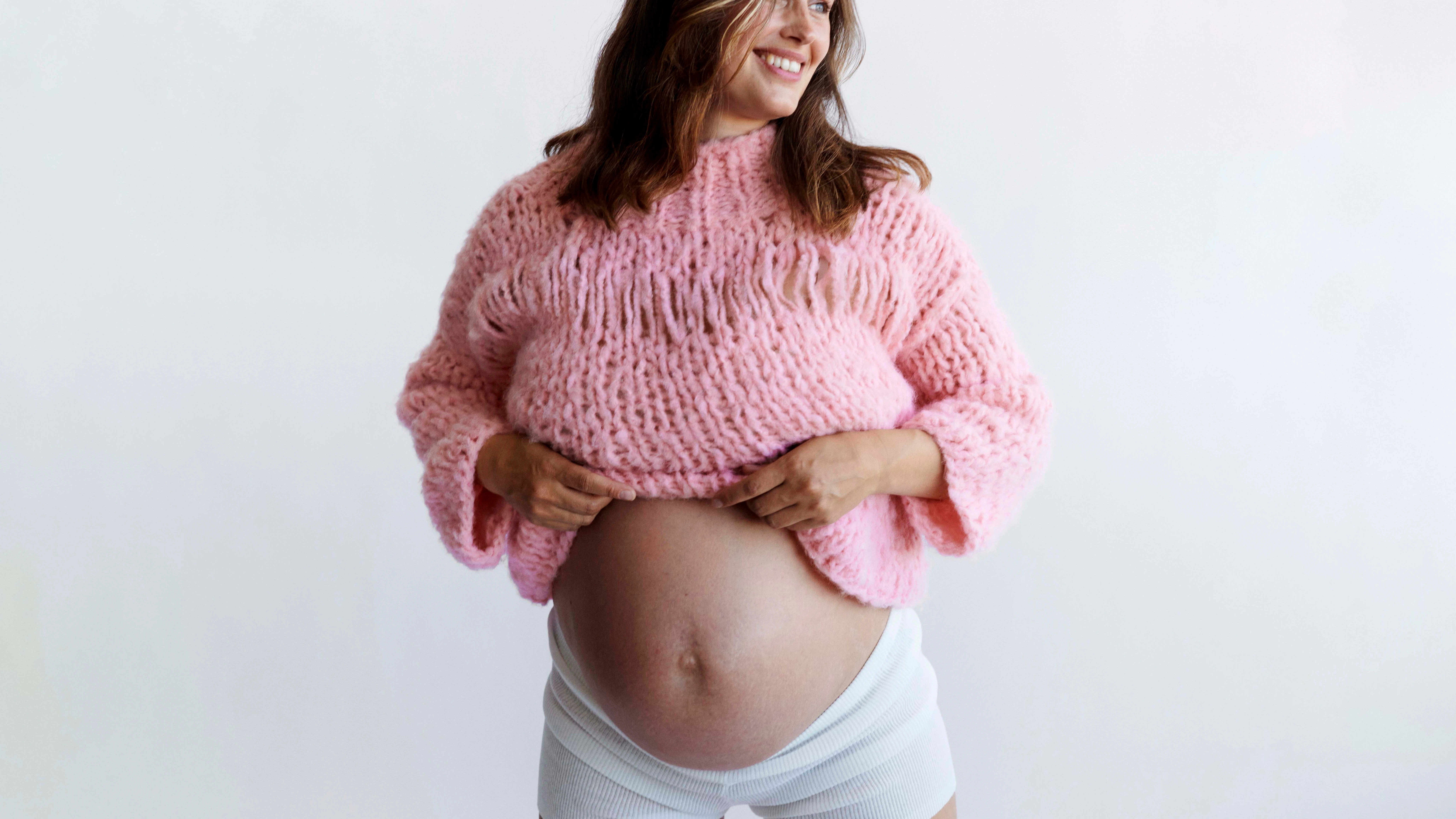 Gravid kvinde smiler og løfter sin lyserøde striktrøje for at vise maven.