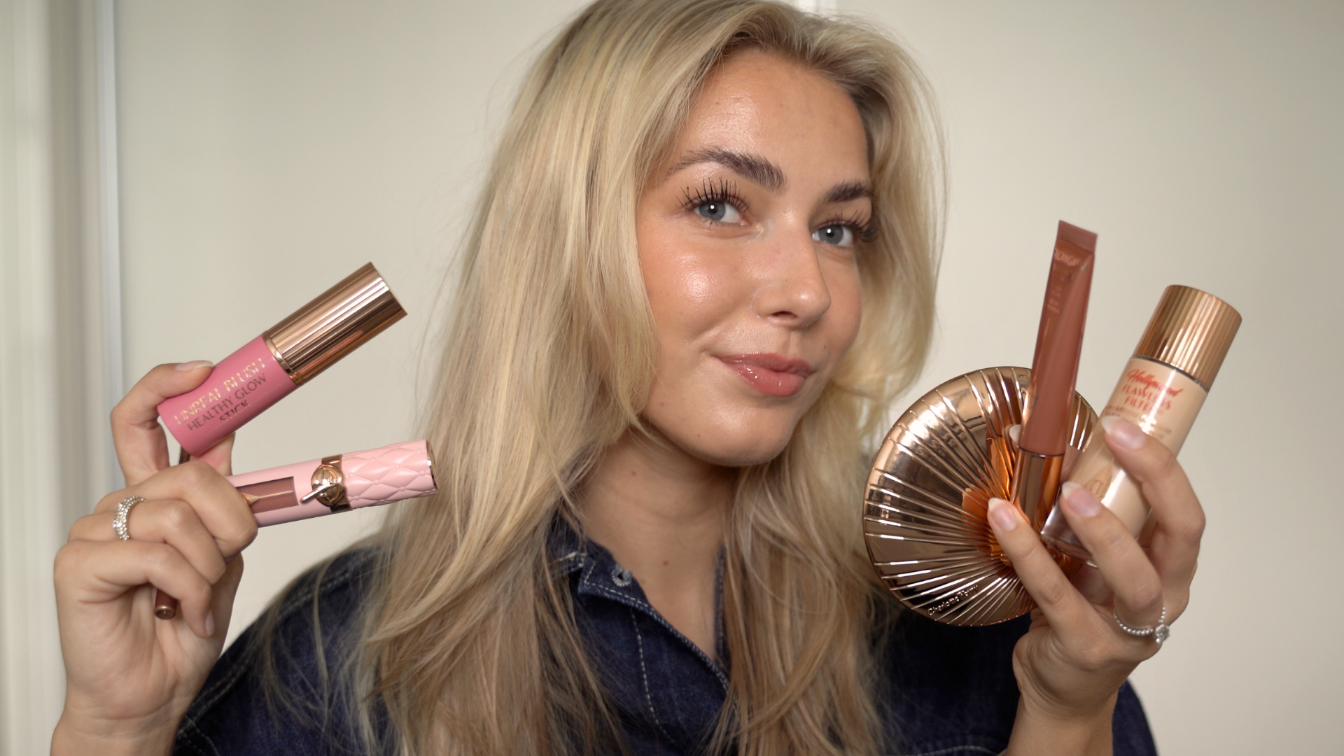 En kvinde holder forskellige makeup-produkter op, herunder en blush stick, læbeglans, pudder og foundation.