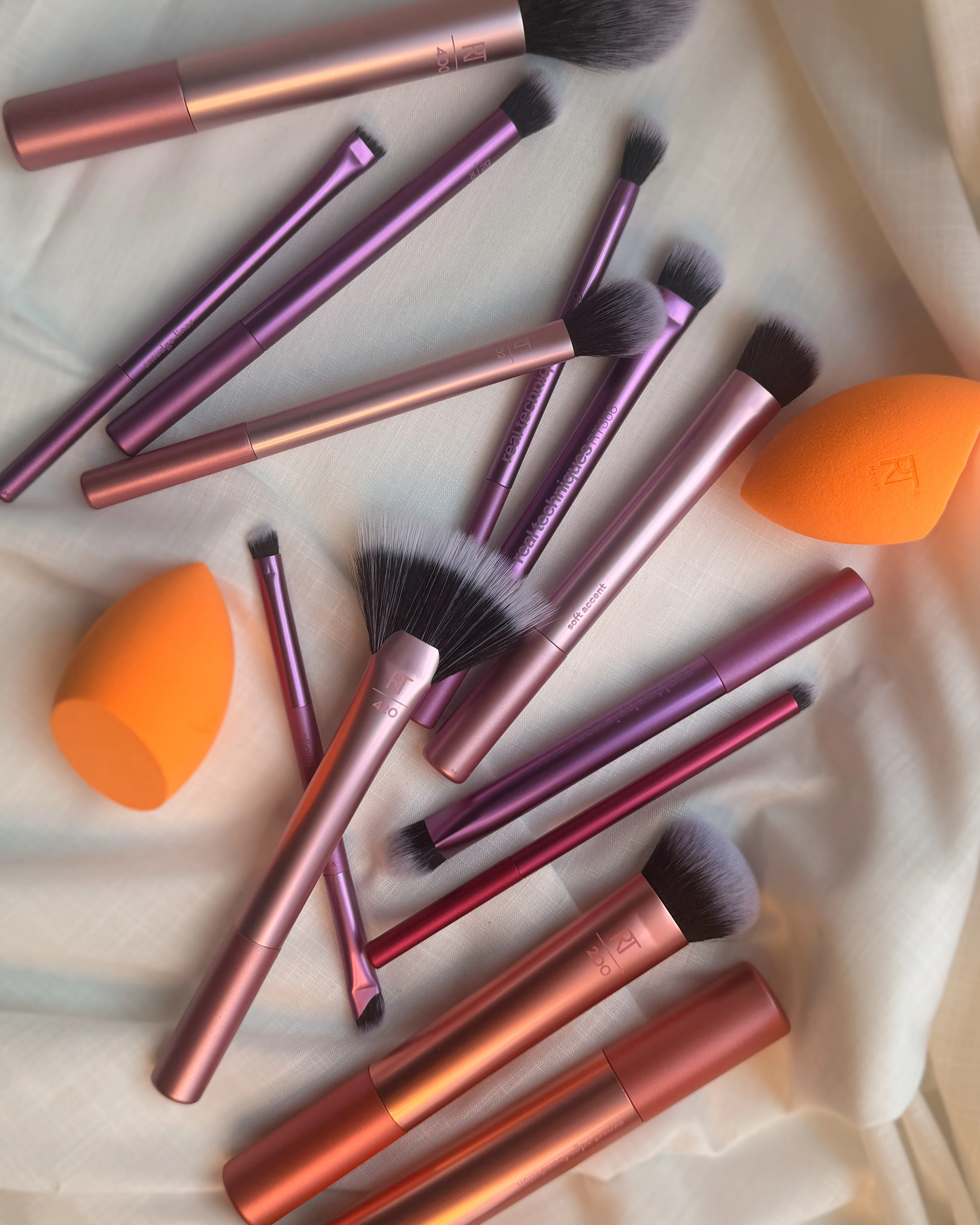 Et udvalg af makeupbørster i rosa og lilla samt orange makeupsvampe spredt på et lyst stof.