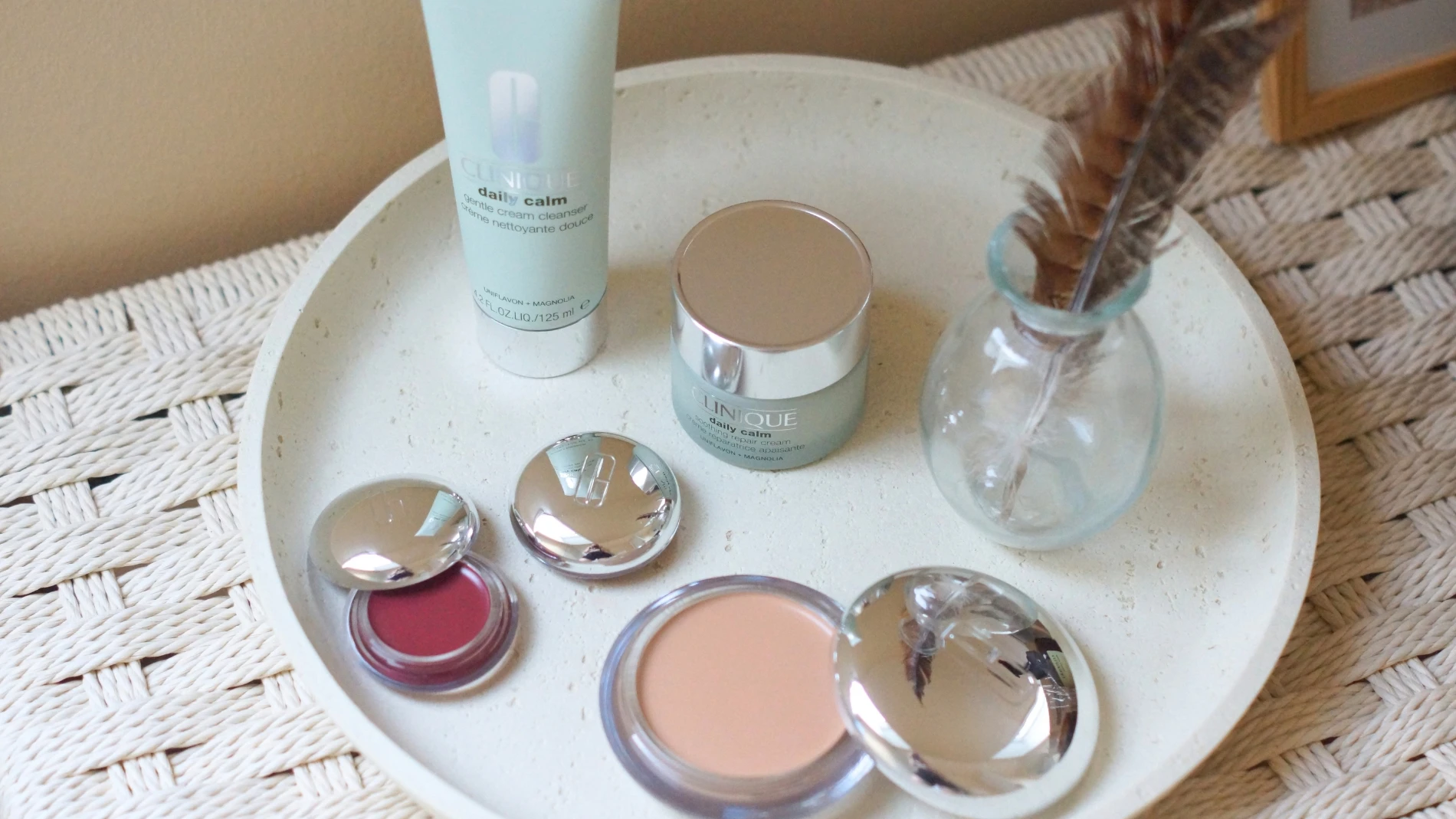Clinique Daily Calm Gentle Cream Cleanser og Soothing Repair Cream, rød læbepomade og foundation på en tekstureret bakke.