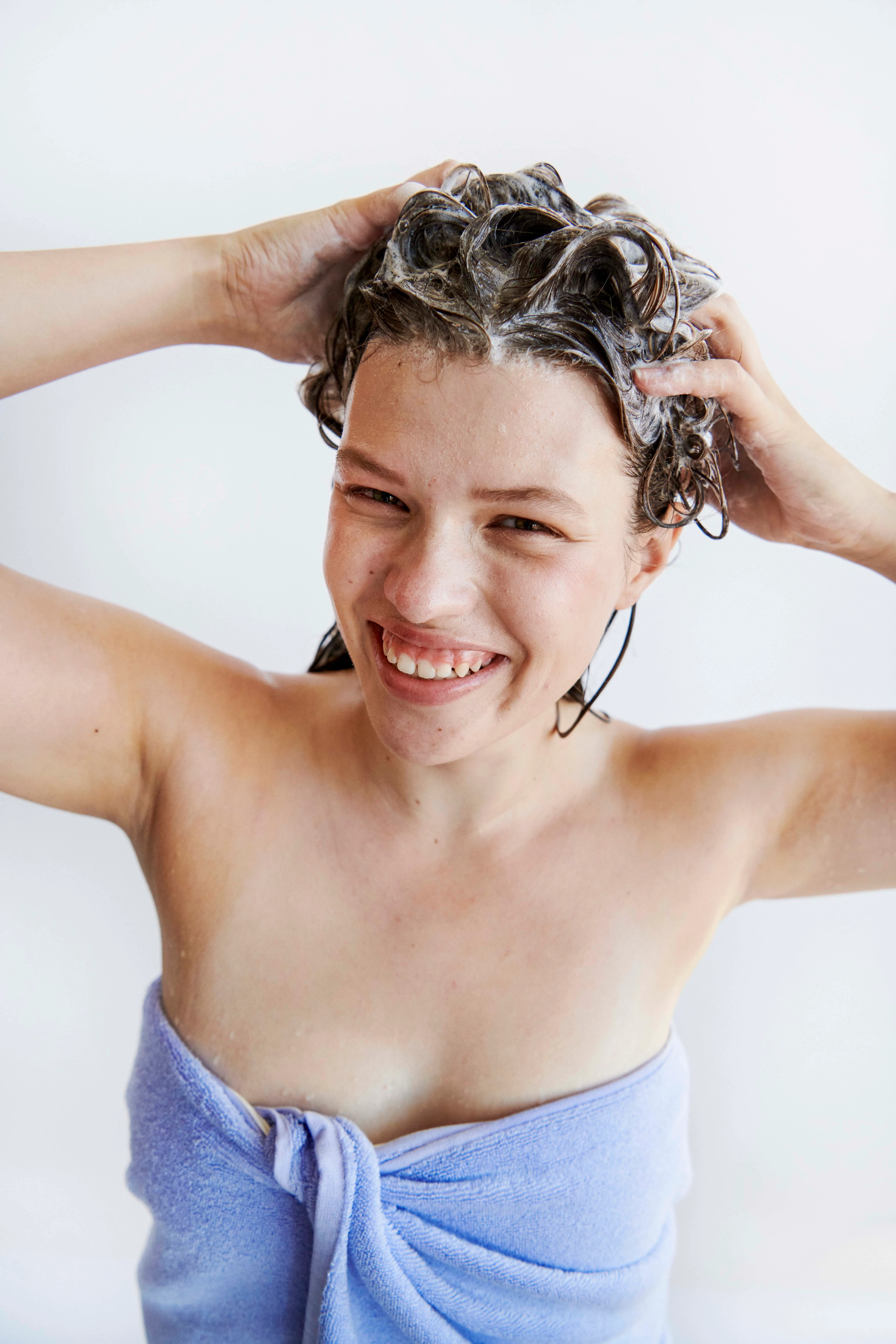 Smilende person vasker vådt, krøllet hår med shampoo og er pakket ind i et lilla håndklæde.