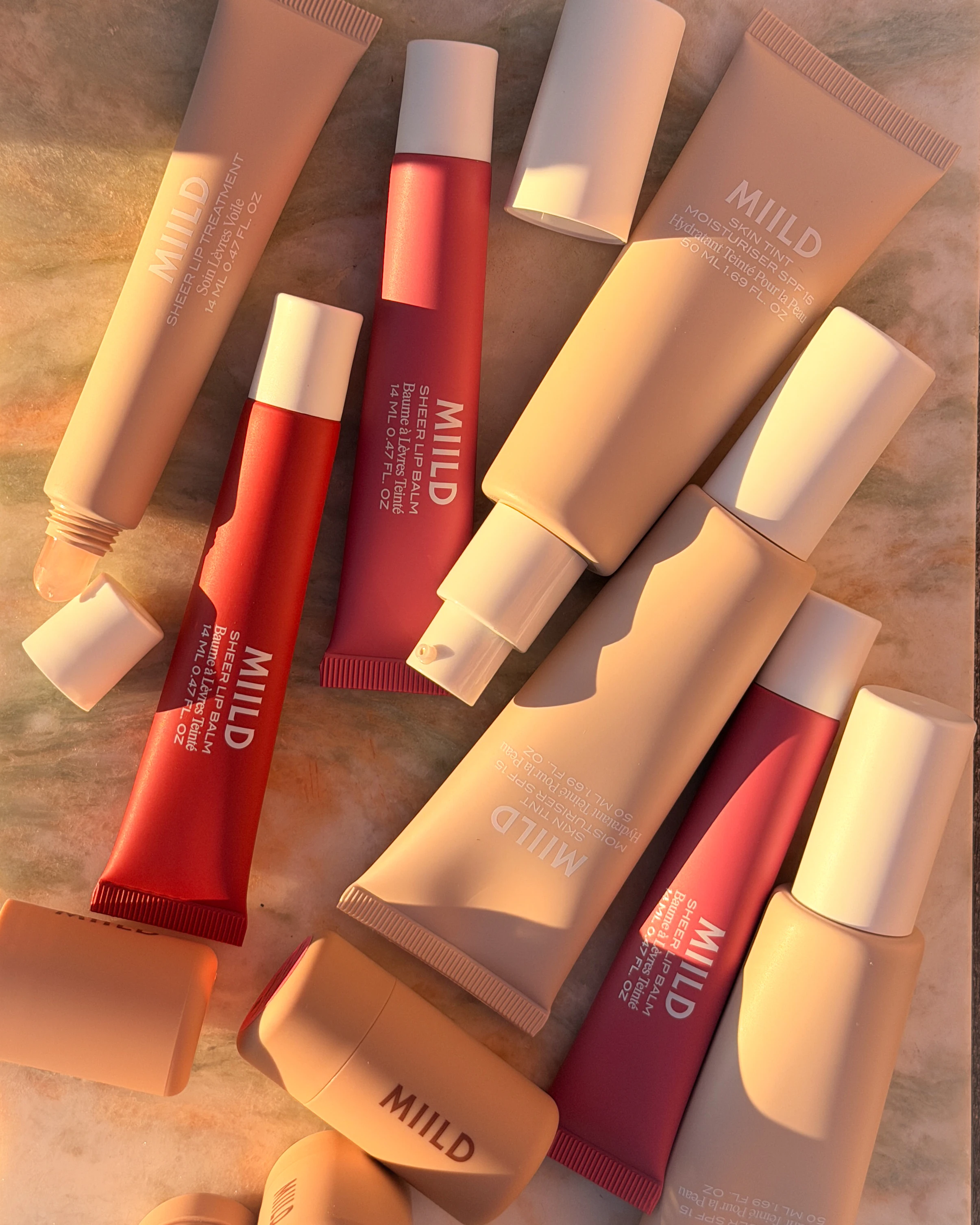 Forskellige MIILD produkter: Skin Tint, Sheer Lip Treatment og Sheer Lip Balm i beige og rød.