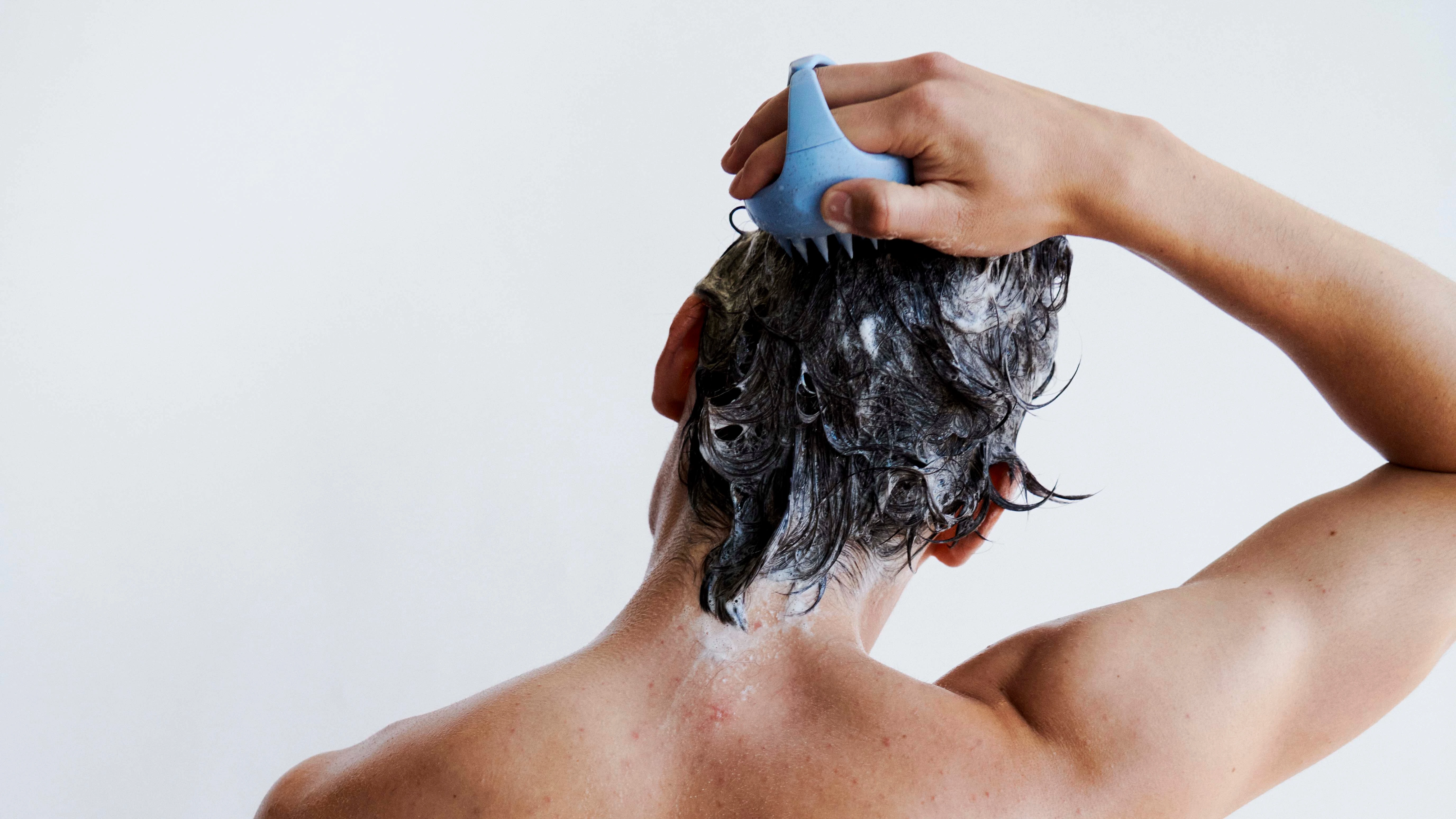 Person set bagfra vasker hår med shampoo og en blå hovedbundsmassager, iført et stribet håndklæde.