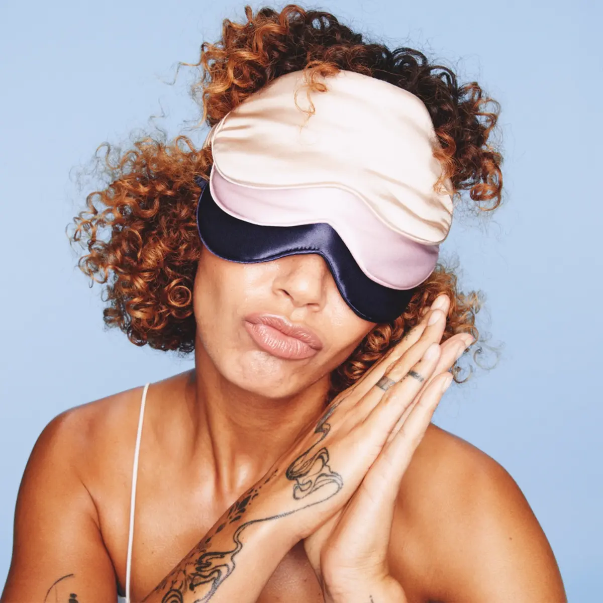 Person med lockigt hår bär två sidenfärgade sovmasker i rosa och mörkblått framför ljusblå bakgrund. Fokus på sleep mask och nattaccessoar för skönhetssömn och self care.