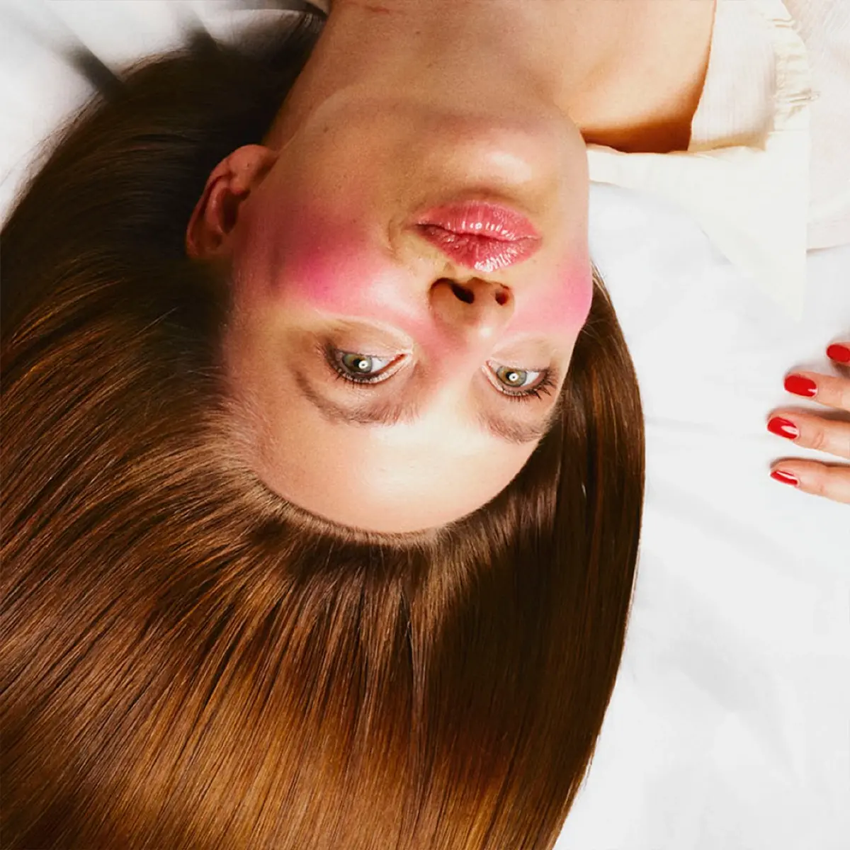 Person med langt, glansfullt brunt hår ligger mot en lys bakgrunn. Makeup-looken fremhever rosa blush på kinnene, naturlig hudfinish og glansfulle lepper, mens håret er spredt ut for å vise hårpleie og glans.