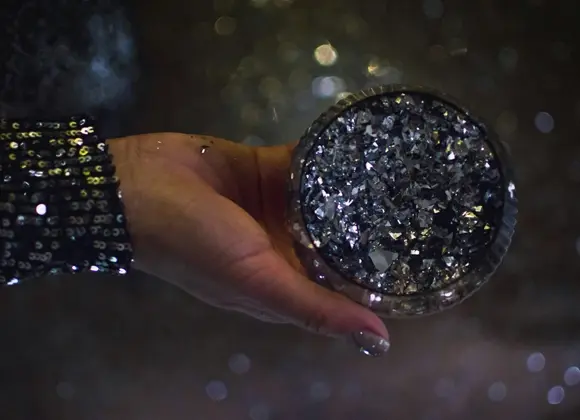 Käsi, jossa on lakatut kynnet ja tumma, kimaltava hiha, pitelee pyöreää, tummalla glitterillä päällystettyä rasiaa hämärää ja sumeaa taustaa vasten.