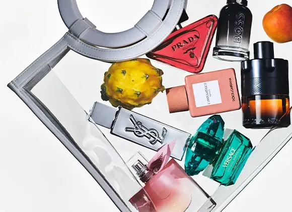 Flera parfymflaskor arrangerade i en transparent handväska tillsammans med frukt. Synliga parfymflaskor från Prada, BOSS, Yves Saint Laurent, Dolce & Gabbana, Versace och Lancôme med olika färgade glasflaskor.