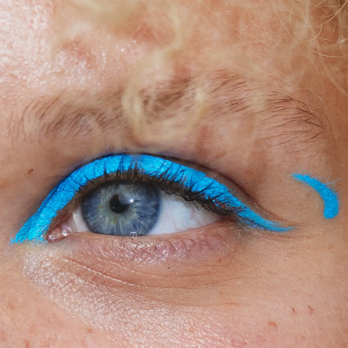 Lähikuva silmistä, joiden yläluomilla on intensiivisen turkoosinsininen eyeliner, joka jatkuu graafisina siipinä silmäkulmissa. Molempien silmien ulkopuolella näkyy pieniä turkoosinsinisiä yksityiskohtia.