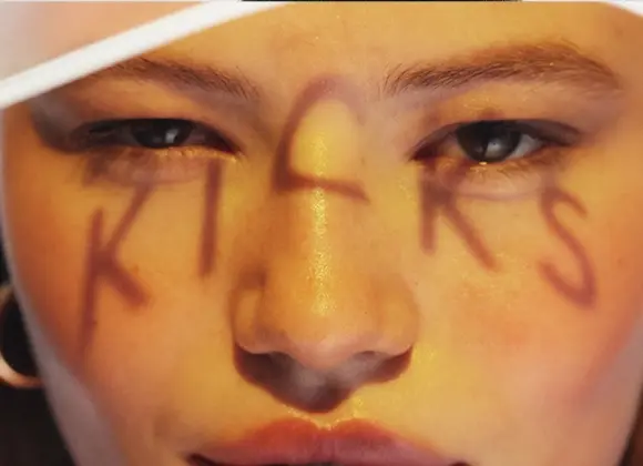 Närbild av ansikte med varm, gyllene makeup och lyster på huden, där bokstäverna "KICKS" är målade över ansiktet. Fokus på ögon, hudfinish och kreativ makeuplook.