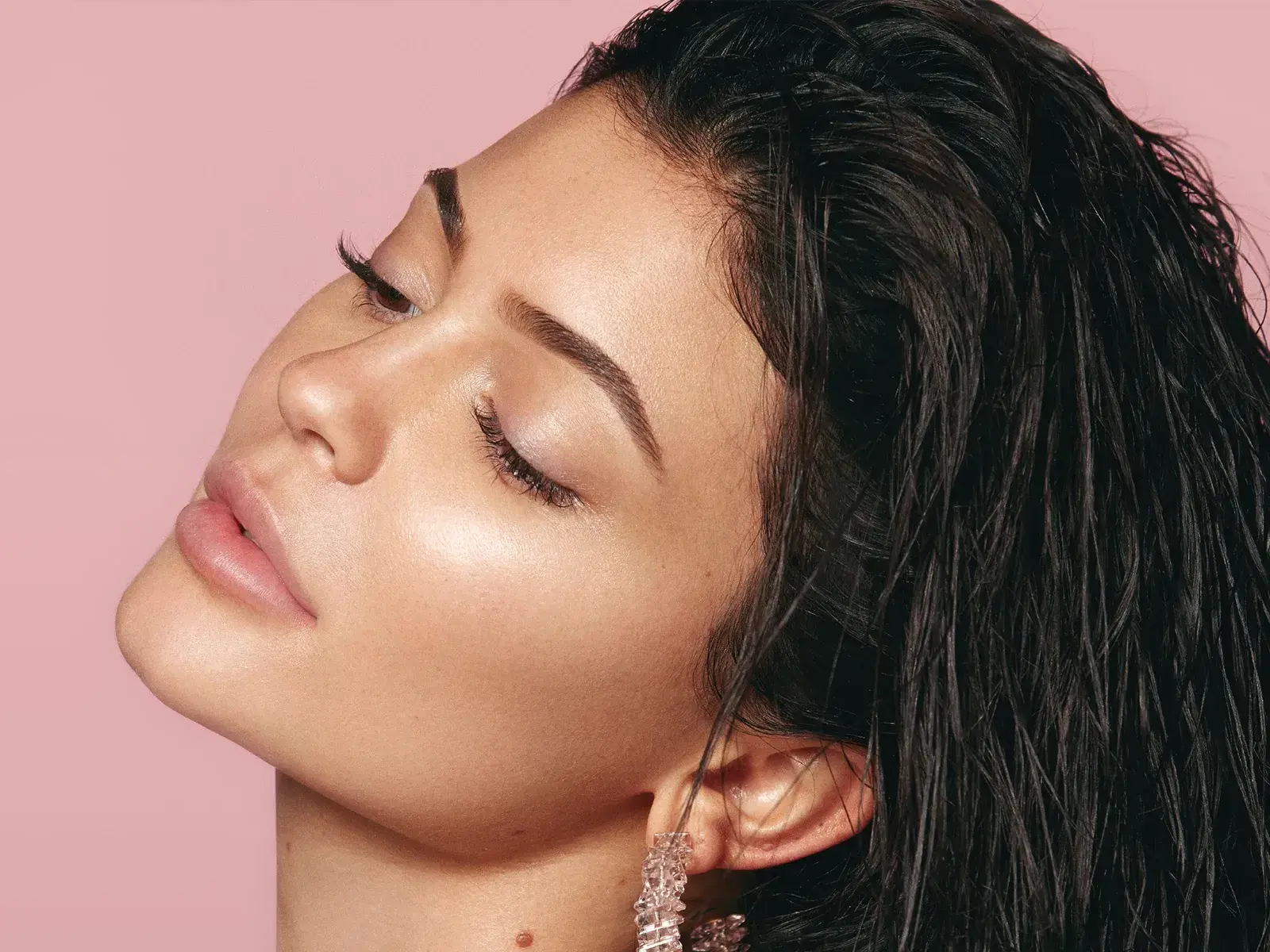 Kylie Jenner profiilissa, katsoo alas ja kallistaa päätään taaksepäin vaaleanpunaista taustaa vasten.