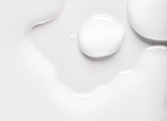 Närbild av vit hudvårdsprodukt med droppar och mjuk, glansig serum- eller lotiontextur mot vit bakgrund. Den krämiga konsistensen framhäver en återfuktande skincare-produkt med lätt och silkeslen finish.