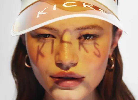 Närbild av en person med solkysst makeup och gyllene highlighter på näsan, med ordet “KICKS” skrivet över ansiktet. Personen bär en vit solskärm med texten “KICKS”, vilket ger en somrig känsla kopplad till makeup och solskydd.