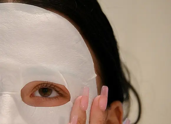 Lähikuva henkilöstä, jolla on valkoinen sheetmask kasvoilla. Henkilöllä on tummat hiukset ja vaaleanpunaiset kynnet. Tausta on beige.