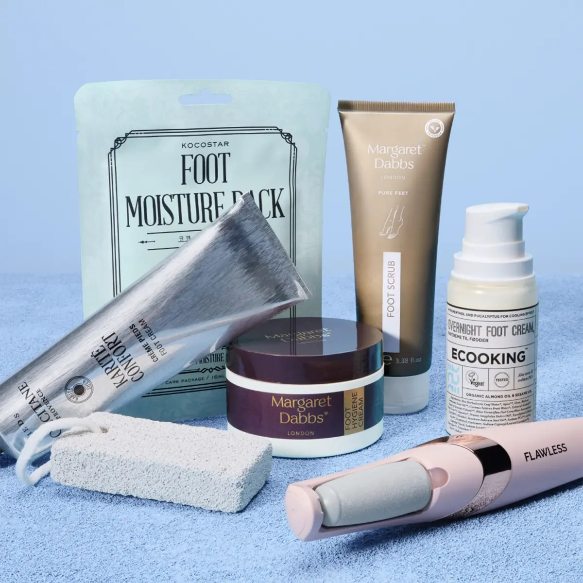 Fotpleieprodukter arrangert mot lys blå bakgrunn med fotmasken KOCOSTAR Foot Moisture Pack, Margaret Dabbs Foot Scrub og Intensive Hydrating Foot Lotion samt COOKING Underfoot Foot Cream. Bildet viser også en elektrisk fotfil fra Flawless, en fotkrem i sølvtube og en hvit pimpstein for eksfolierende og mykgjørende fotpleie hjemme.