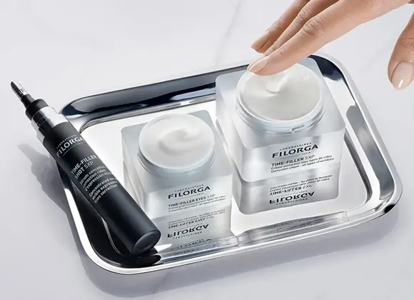 En hånd berører en krukke med Filorga "TIME-FILLER 5XP" krem, som står på et sølvbrett sammen med en mindre krukke og en tube med "TIME-FILLER EYES 5XP".