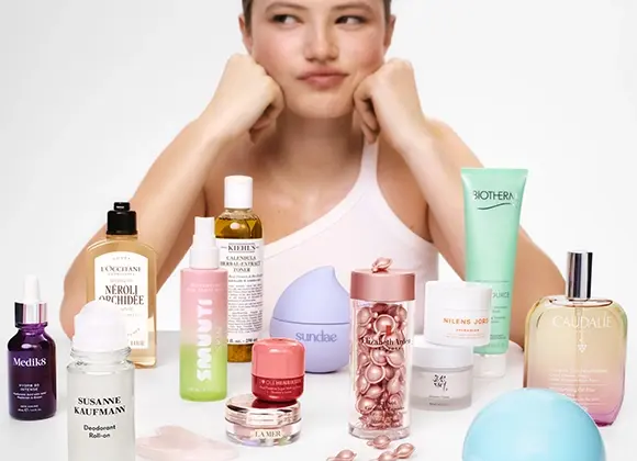 Henkilö nojaa leukaa käsiinsä pöydän takana, joka on täynnä ihonhoito- ja kauneustuotteita, kuten L’Occitane Néroli & Orchidée, Kiehl’s Calendula Herbal Extract Toner, Biotherm Aquasource, NUXE Huile Prodigieuse, Caudalie Eau des Vignes, Medik8 -seerumi, Susanne Kaufmann Deodorant Roll-On ja Elizabeth Arden -kapseliseerumi.