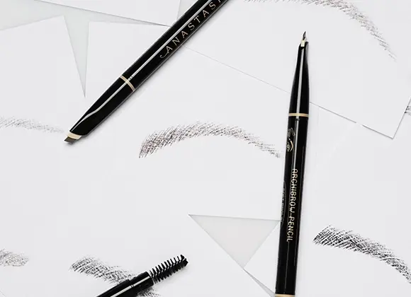 Svart ögonbrynspenna från Anastasia Beverly Hills med texten "ANASTASIA" och "BROW WIZ", tillsammans med en spoolieborste och flera streck av brynfärg på vit bakgrund.