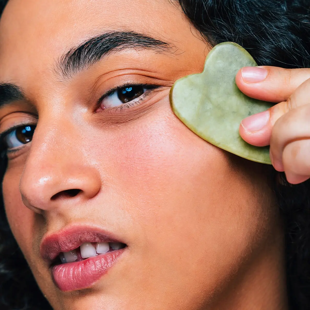 Nærbilde av en person som bruker en grønn gua sha-stein mot kinnet som del av en hudpleierutine. Ansiktet, hånden og massasjeverktøyet er tydelig synlige mot en nøytral bakgrunn, med fokus på skincare og ansiktsmassasje.