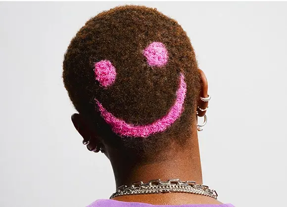 Baksidan av ett huvud med kort, mörkt hår där en rosa smileyform är skapad i håret. Personen bär flera örhängen och halskedjor mot en ljus bakgrund.