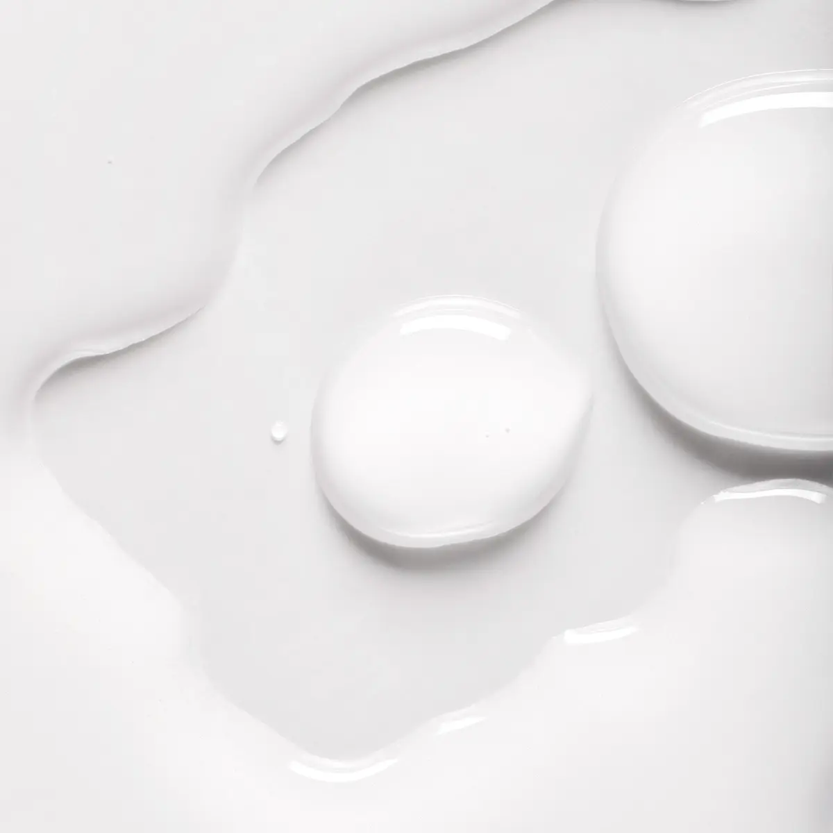 Lähikuva valkoisesta ihonhoitotuotteesta, jossa näkyy tippoja ja pehmeä, kiiltävä seerumi- tai lotion-koostumus valkoista taustaa vasten. Voidemainen rakenne korostaa kosteuttavaa skincare-tuotetta, jossa on kevyt ja silkkinen lopputulos.
