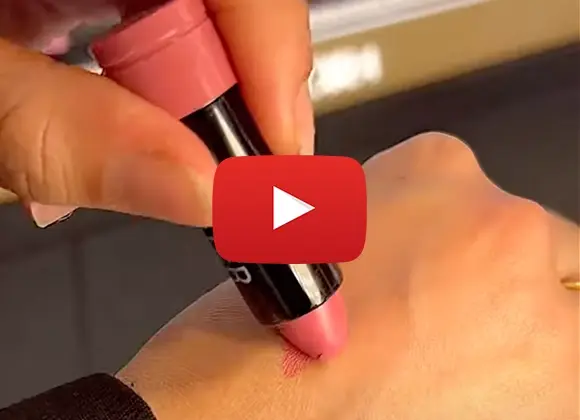  En hand applicerar en rosa läppstift på en annan hand för att visa färgen. Ett Youtube play-tecken är placerat i mitten av bilden.