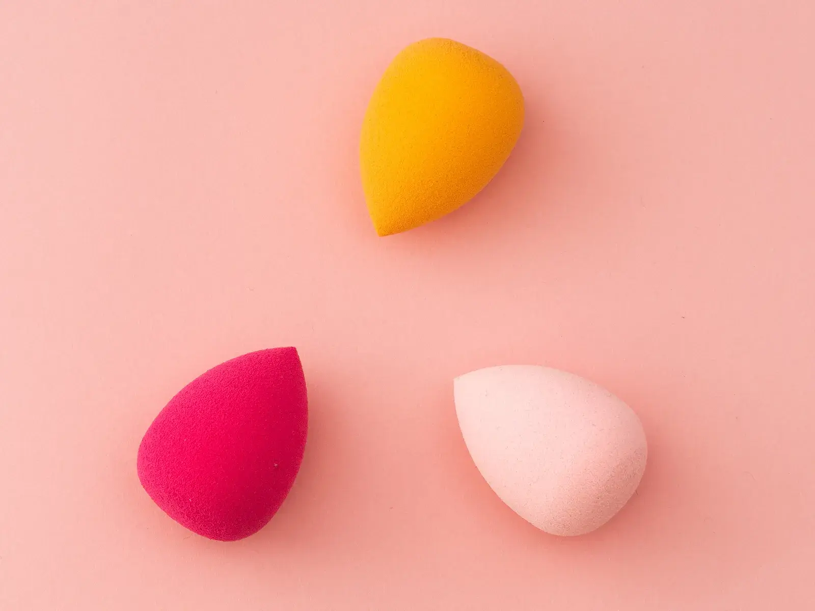 Tre sminksvampar i olika färger (gul, rosa och röd) på en rosa bakgrund.