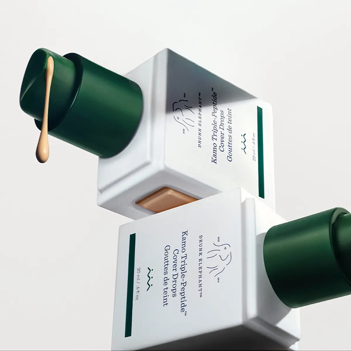 Två flaskor Drunk Elephant Kamo Triple-Peptide Cover Drops, där en doserar ut en droppe foundation. Text: 20 ml / .6 fl oz.