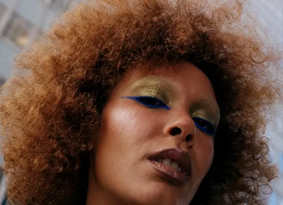Nærbilde av en person med fyldig krøllete hår og makeup med gullfarget øyenskygge og markert blå eyeliner, samt naturlig base og nude lepper.