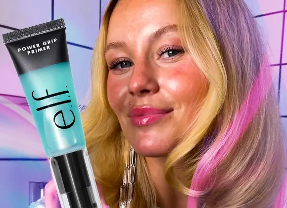 Närbild av en person med långt hår som håller upp en tub med texten ”e.l.f. Power Grip Primer”. Personen är fotograferad framifrån mot en färgstark bakgrund med grafiska linjer i rosa och blå toner. Produkten har en genomskinlig turkos tub med svart kork och synlig logotyp.
