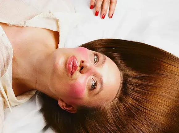 En person ligger mot en hvit bakgrunn med langt, rett brunt hår spredt rundt hodet. Kinnene er markert med sterkt rosa rouge, og leppene har en blank rosafarget glans. En hånd med rødlakkerte negler vises over hodet.