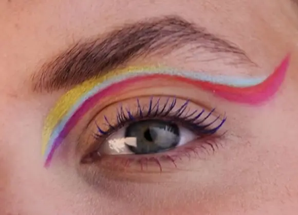 Lähikuva silmästä, jossa on monivärinen eyeliner yläluomella, sävyinä keltainen, vaaleanpunainen ja vaaleansininen. Ripset ovat korostetut ja tausta on neutraali.