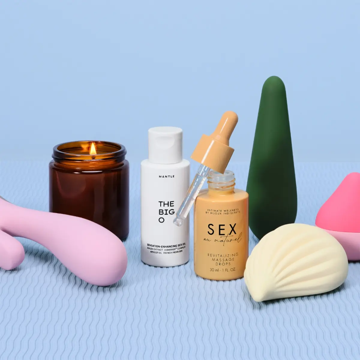 Stillbild med intimprodukter och kroppsvård på ljusblå bakgrund. Ett tänt doftljus står bredvid Mantle The Big S*xcation Enhancing Serum och en flaska “Sex Oil” revitalizing massage oil med pipett, tillsammans med rosa och gröna sexleksaker.