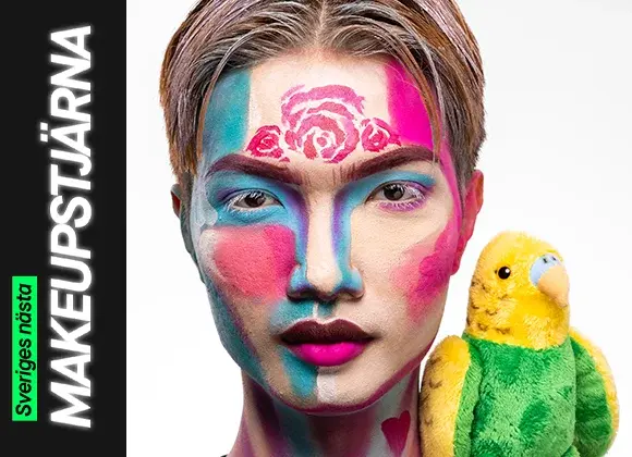 En person med färgstark ansiktsmålning i blå, rosa och lila blockfärger tillsammans med målade rosor i pannan och neonrosa läppar, poserar bredvid en grön och gul gosedjursfågel.