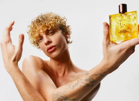 Person med kort lockigt hår håller upp en gul parfymflaska med texten "Versace Eros Energy" och Versace-logotypen framtill. Flaskan är transparent med gyllene vätska och svart kork mot en ljus bakgrund.
