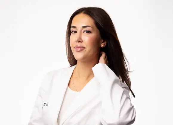 Porträtt av en person med långt, mörkt hår som bär en vit kavaj och en ljus topp. Personen står mot en vit bakgrund och håller ena handen bakom nacken.