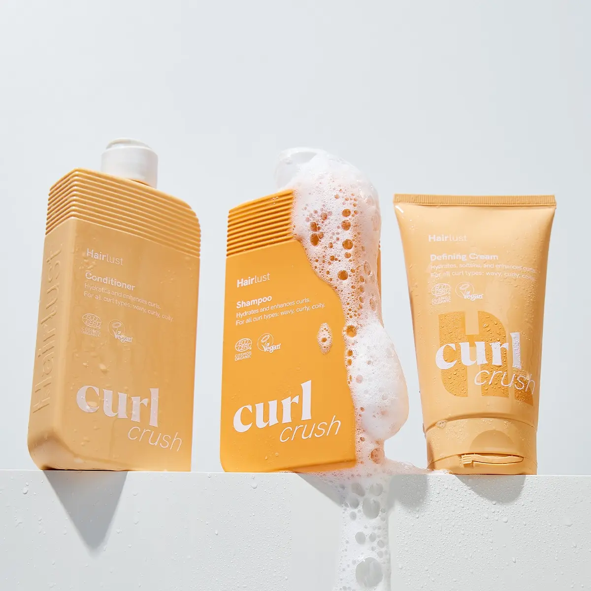 Kolme Hairlust Curl Crush -tuotetta kiharille hiuksille valkoisella alustalla: Conditioning Wash, Shampoo ja Defining Crème. Pakkaukset ovat beigen ja oranssin sävyisiä, ja valkoista vaahtoa valuu shampoopullon yli. Näkyvä teksti sisältää sanat ”Hairlust”, ”Conditioner Wash”, ”Shampoo”, ”Defining Crème”, ”Vegan” ja ”curl crush”.