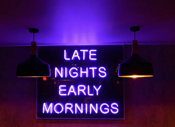 Neonljusskylt med texten "Late Nights Early Mornings" i ett mörkt rum, upplyst av två hängande lampor
