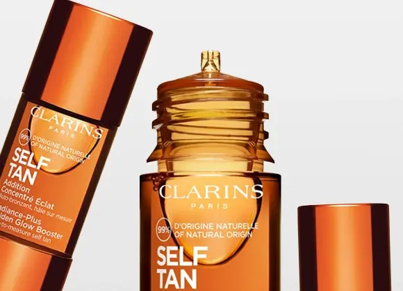 Lähikuva ruskeansävyisistä Clarins self tan -tuotteista, joissa lukee “CLARINS PARIS”, “SELF TAN” ja “99% D’ORIGINE NATURELLE / OF NATURAL ORIGIN” vaaleaa taustaa vasten. Kiiltävän kuparinväriset pakkaukset esittelevät itseruskettavia tuotteita kasvoille ja vartalolle.