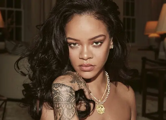 Rihanna tittar in i kameran iklädd halsband och visar hand med tatueringar. Sitter i vardagsrumsmiljö.