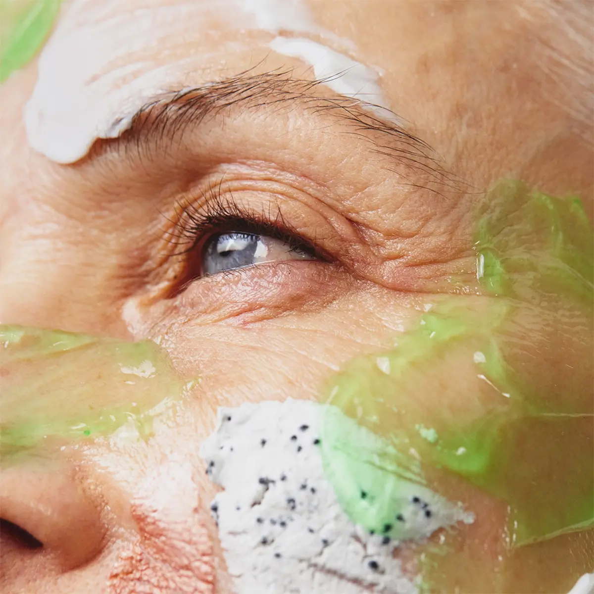 Närbild av ett öga med synliga hudvårdsprodukter applicerade runt ögat och på kinden. En grön gelmask och en vit exfolierande ansiktsmask med svarta korn framhäver hudvård och ansiktsbehandling.