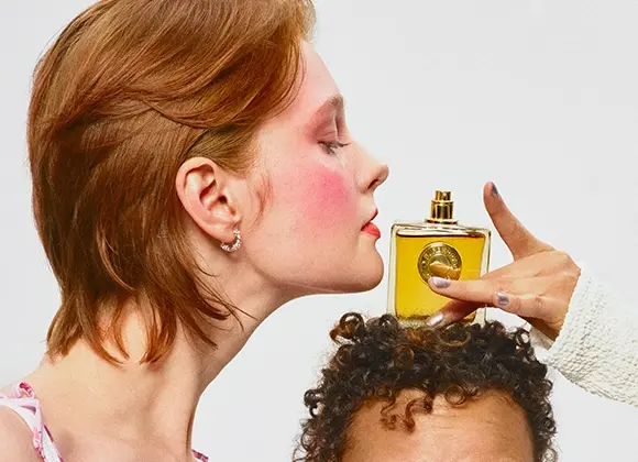 En person lutar sig fram och doftar på en gul parfymflaska som hålls ovanför en annan persons lockiga hår. Närbild av parfymflaska med gul vätska och rund guldetikett, med makeup som rouge och rött läppstift synligt.