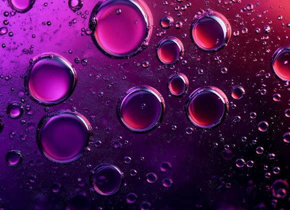 Lähikuva violetinvärisistä kuplista vedessä, taustalla tummia sävyjä ja häivähdyksiä vaaleanpunaisesta ja sinisestä.