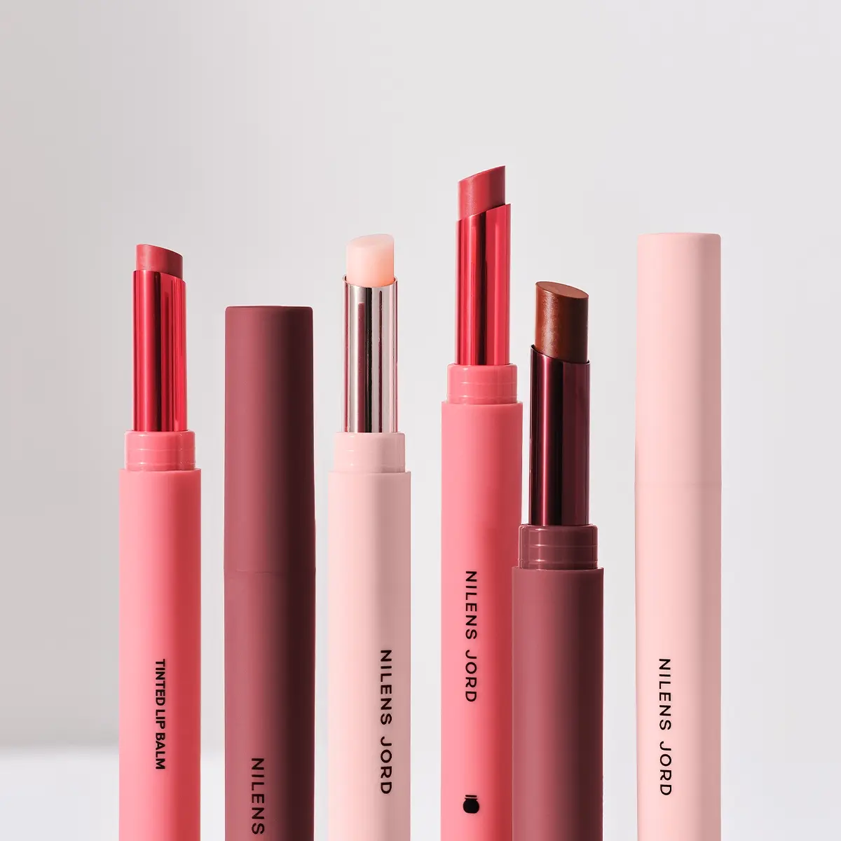 Fem leppebalmer fra Nilens Jord i rosa og vinrød emballasje mot lys bakgrunn, med synlig tekst "TINTED LIP BALM", "NILENS" og "NILENS JORD". Leppebalmene vises i rosa, transparent lys og brun nyanse.