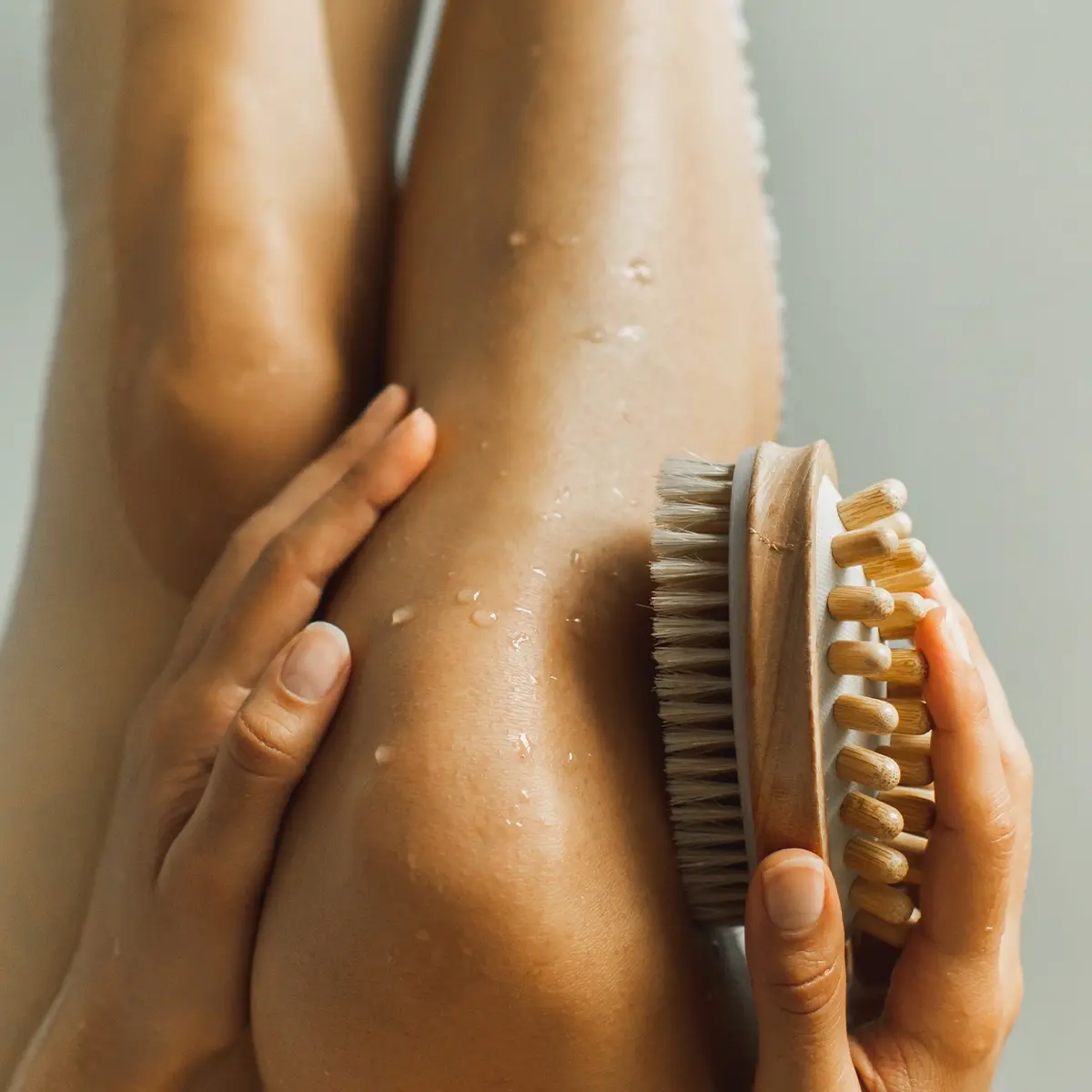 Henkilö käyttää kuivaharjaa vartalonhoitoon jalalla, jonka iholla näkyy vesipisaroita. Puinen harja luonnonvärisillä harjaksilla pidetään jalkaa vasten minimalistisessa spa- ja wellness-ympäristössä, jossa korostuvat kuorinta ja body care.