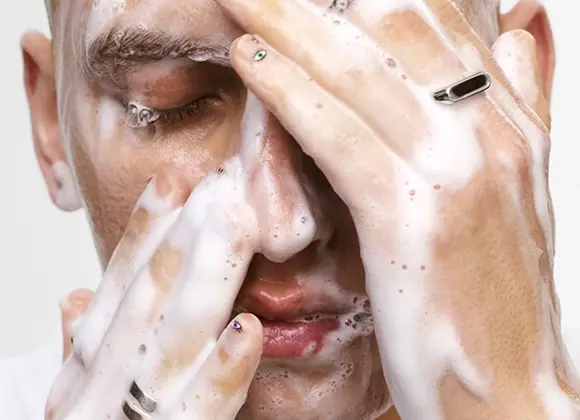 Lähikuva henkilöstä, joka puhdistaa kasvojaan vaahtoavalla kasvojenpuhdistusaineella skincare-rutiinissa, kädet peitettynä vaahdolla otsalla, poskilla ja nenällä. Sormessa näkyy hopeanvärinen sormus, kun puhdistusvaahtoa hierotaan ihoon.