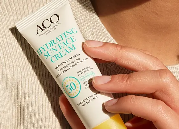Närbild av en hand som håller en tub ACO Hydrating Sun Face Cream SPF 50 med texten “Invisible on skin”, “For everyday use works well under make-up”, “High protection 50” och “For sensitive skin unperfumed”, mot en neutral bakgrund. Fokus på solskydd för ansiktet och hudvård.